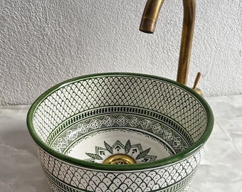 May include: A white ceramic sink with a green and white geometric pattern. The sink has a round shape and a drain in the center. A gold faucet is visible in the background.