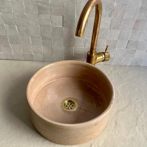 A round, light brown ceramic sink with a gold-colored drain and faucet. The faucet has a curved spout and a single handle. The sink is set against a backdrop of small, square, light gray tiles.
