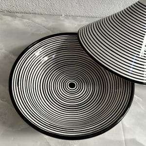 K&ouml;nnte beinhalten: Eine schwarze und wei&szlig;e Keramik-Servierplatte mit einem konzentrischen Kreis-Muster. Die Platte ist rund und hat einen erh&ouml;hten Rand.