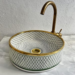 Può includere: Un lavabo in ceramica bianca con un motivo geometrico verde e nero. Il lavabo ha un bordo dorato e un rubinetto in ottone.
