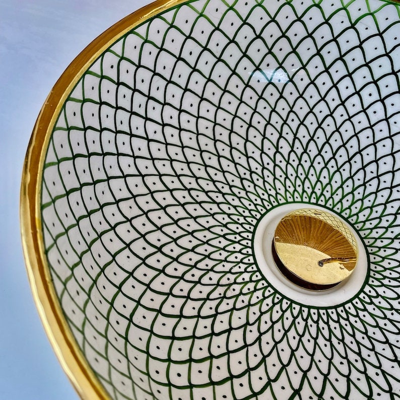 A white ceramic bowl with a gold rim and a green and white geometric pattern. The bowl has a gold-colored drain in the center.