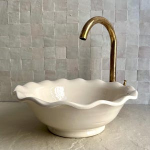 May include: A cream-colored ceramic sink with a scalloped edge, paired with a curved brass faucet. The sink sits on a countertop, with a square tile backsplash in the background. The faucet has a single handle.