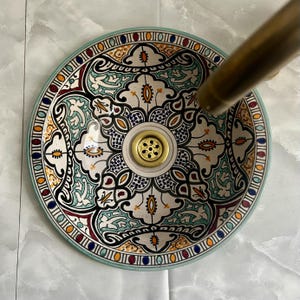 May include: A white ceramic sink with a colorful Moroccan-inspired design. The sink has a round shape and a drain in the center. The design features intricate patterns in shades of blue, green, red, and yellow.