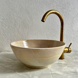Puede incluir: Un lavabo de cerámica blanco con un grifo dorado. El lavabo es redondo y tiene un ligero borde alrededor del borde. El grifo es alto y tiene un caño curvo.