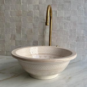 Puede incluir: Un lavabo de cerámica beige con un patrón decorativo y un grifo dorado. El lavabo tiene forma de cuenco con un borde ancho y se apoya en un pequeño pedestal. El grifo es alto y curvado, con una boquilla simple.