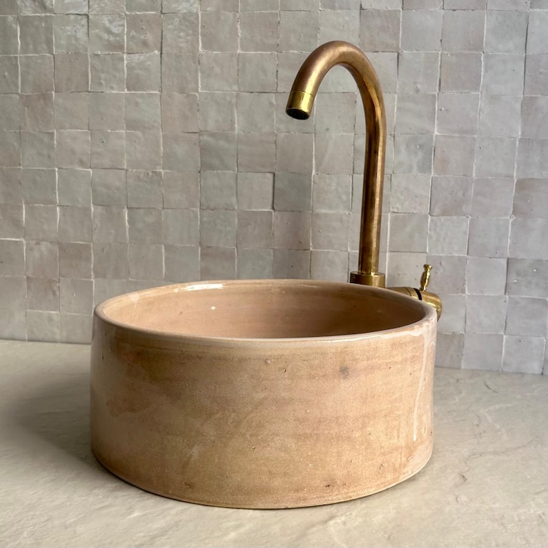 A round, light brown ceramic sink with a brass faucet. The sink has a simple, modern design and is set against a backdrop of square, off-white tiles. The faucet has a curved spout.