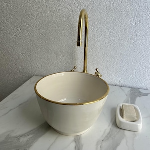 Può includere: Un lavandino in ceramica bianca con un bordo dorato e un rubinetto dorato. Un portasapone bianco con una saponetta è sul bancone.