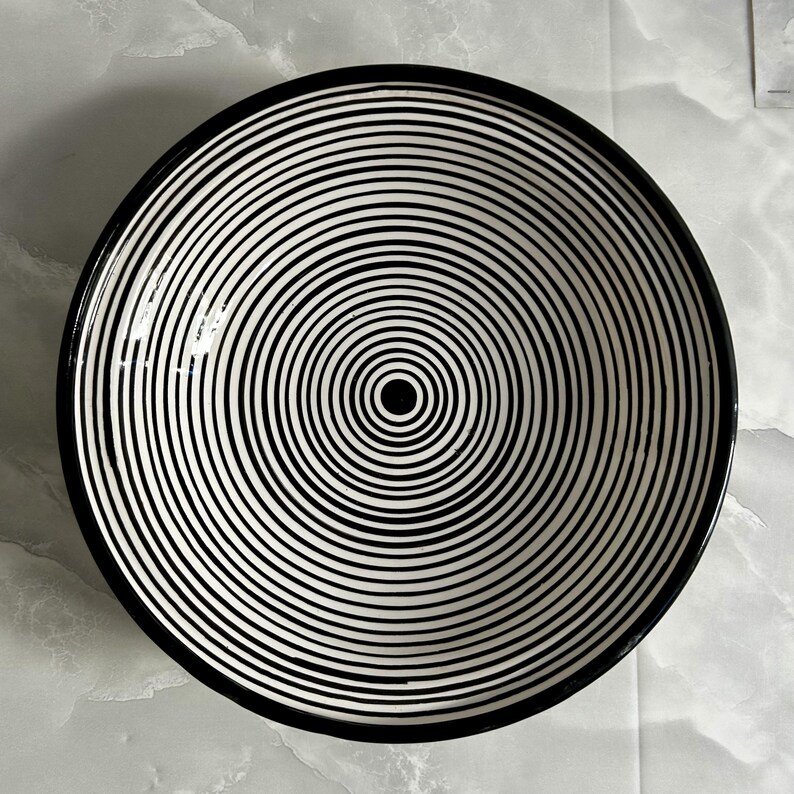 K&ouml;nnte beinhalten: Eine gro&szlig;e, wei&szlig;e Keramiksch&uuml;ssel mit einem schwarzen und wei&szlig;en konzentrischen Kreisdesign. Die Sch&uuml;ssel hat einen schwarzen Rand.