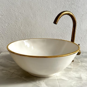 Puede incluir: Lavabo de cerámica blanca con un borde dorado y un grifo de latón.