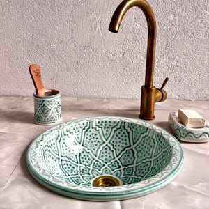 Può includere: Un lavandino in ceramica verde e bianco con intricati motivi geometrici. Il lavandino è rotondo e ha uno scarico in ottone. Un rubinetto dorato è montato sul muro dietro il lavandino.