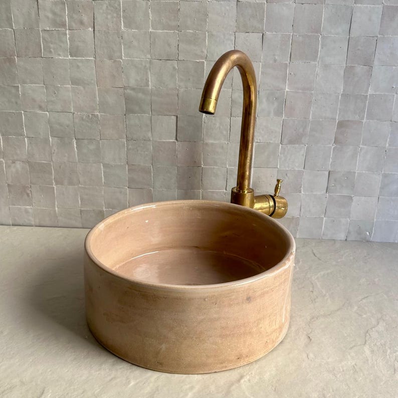 A round, light peach-colored ceramic sink with a brass faucet. The faucet has a curved spout and a single handle. The sink is set against a backdrop of small, square, off-white tiles.