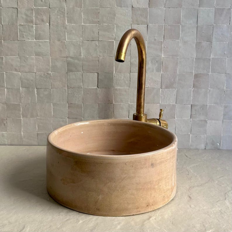 A round, light brown ceramic sink with a brass faucet. The sink has a simple, minimalist design. The faucet is curved and has a single handle. The background is a light-colored tile wall.