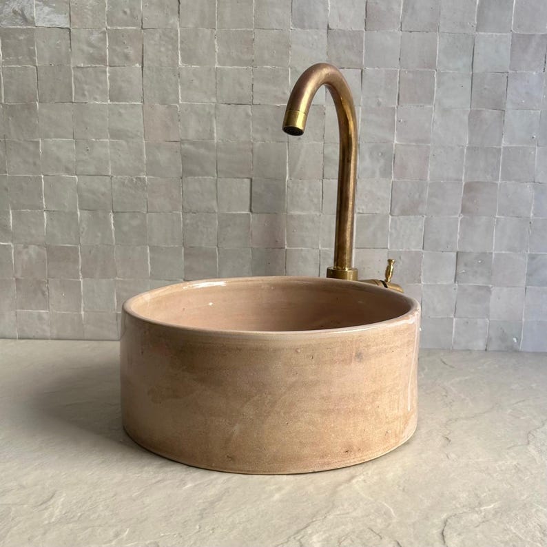 A round, light brown ceramic sink with a brass faucet. The sink has a simple, modern design and is set against a backdrop of small, square, white tiles. The faucet has a curved spout.