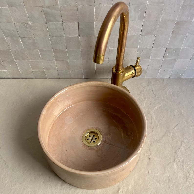 A round, peach-colored ceramic sink with a brass faucet. The faucet has a curved spout and a single handle. The sink has a brass drain and sits on a light-colored surface. The background features a tiled wall.
