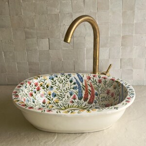 May include: Oval ceramic sink with a floral design in red, blue, and yellow. The sink has a white base and is paired with a gold-colored faucet. The floral pattern covers the interior of the sink, creating a decorative bathroom fixture.