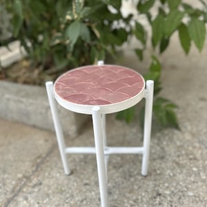 May include: A white metal plant stand with a round, pink tiled top. The stand has a simple, modern design and is perfect for displaying small plants or decorative items.