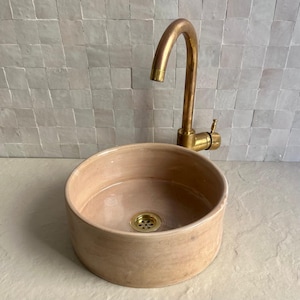 A round, light brown ceramic sink with a brass faucet. The sink has a gold drain and is set against a light-colored tiled wall. The faucet has a curved spout and a single handle.