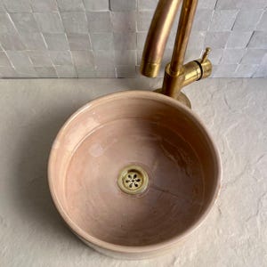 A round, blush-colored ceramic sink with a brass faucet. The sink has a gold-colored drain cover. The faucet is mounted on a white tiled wall. The sink is a vessel sink, designed to sit on top of a counter.