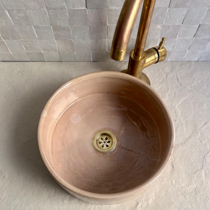 A round, blush-colored ceramic sink with a brass faucet. The sink has a gold-colored drain cover. The faucet is mounted on a white tiled wall. The sink is a vessel sink, designed to sit on top of a counter.
