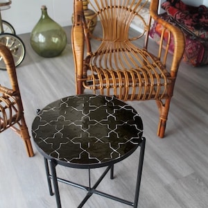 Pode incluir: Uma mesa lateral redonda com tampo de mosaico preto e pernas de metal preto. Uma cadeira de vime está atrás da mesa. Uma garrafa de vidro verde grande está no fundo. A mesa é uma peça de mobiliário elegante.