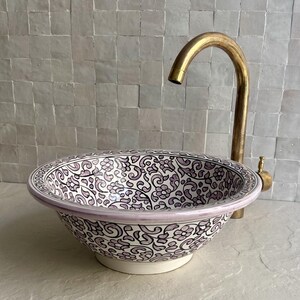 Pode incluir: Uma pia de cerâmica decorativa com um padrão floral em branco, preto e lavanda. A pia tem formato arredondado e uma borda lavanda clara. Uma torneira dourada é instalada acima da pia.