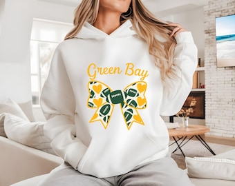 May include: White hoodie with the text "Green Bay" in yellow script. A large bow design features green footballs and yellow hearts. The hoodie is a comfortable, casual style.