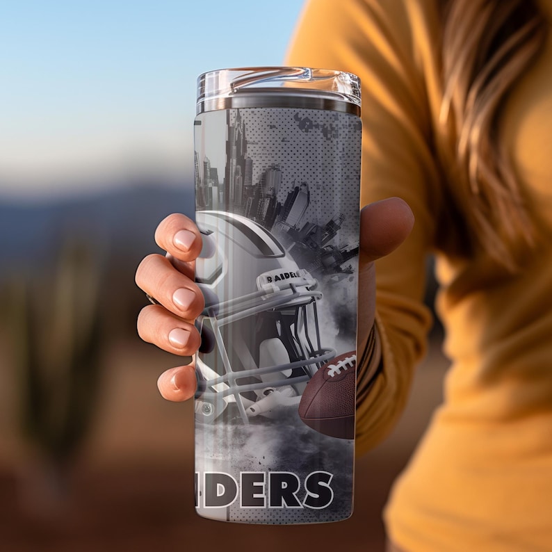 May include: A stainless steel tumbler with a black and white graphic of a football helmet and football with the word "RAIDERS" printed on the bottom. The graphic is set against a cityscape background.