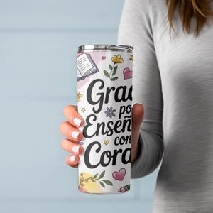 Pu&ograve; includere: Un bicchiere in acciaio inossidabile bianco con un design floreale rosa, giallo e verde. Il bicchiere ha il testo "Gracias por ense&ntilde;ar con coraz&oacute;n" in nero.