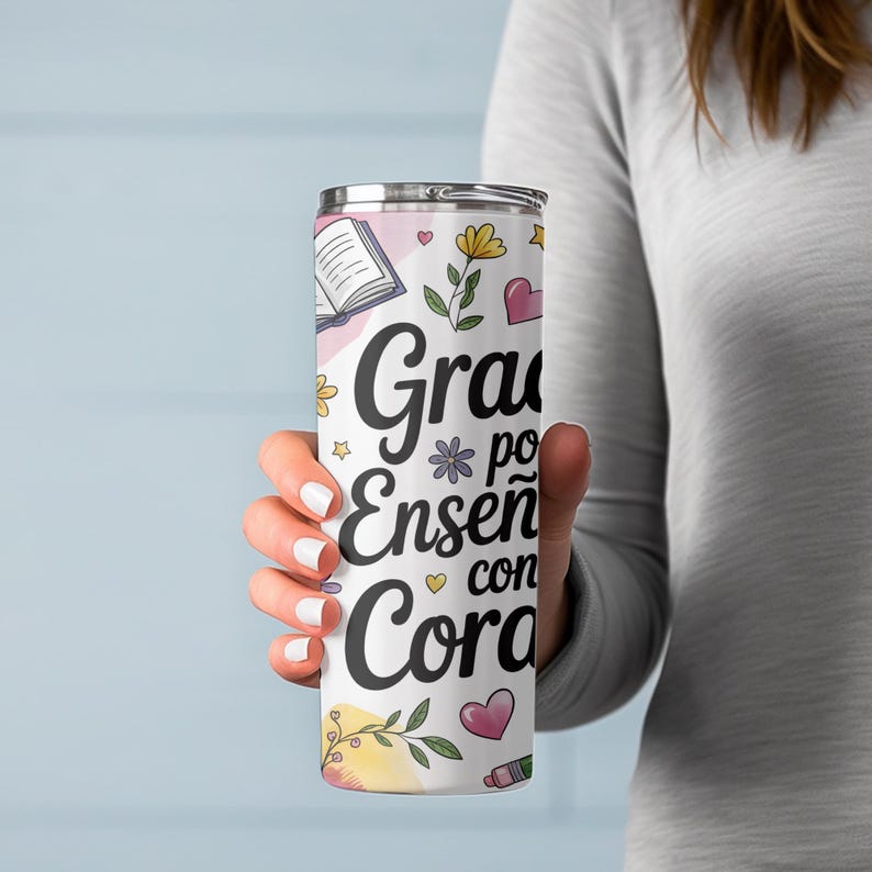 Pu&ograve; includere: Un bicchiere in acciaio inossidabile bianco con un design floreale rosa, giallo e verde. Il bicchiere ha il testo "Gracias por ense&ntilde;ar con coraz&oacute;n" in nero.