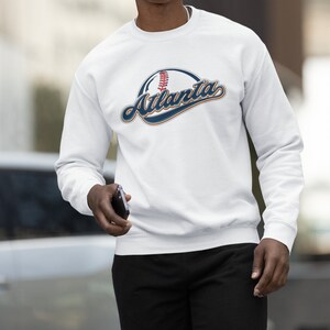 May include: White long-sleeved sweatshirt with a navy blue and orange graphic of a baseball with the word "Atlanta" in a stylised font.