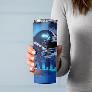May include: A blue and silver insulated tumbler featuring a football helmet design. The tumbler has a silver lid and the word "ALL" in large blue letters. The design is set against a smoky blue background.