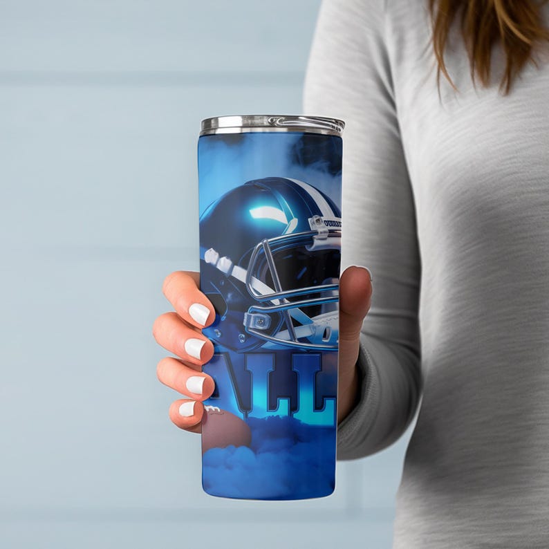 May include: A blue and silver insulated tumbler featuring a football helmet design. The tumbler has a silver lid and the word "ALL" in large blue letters. The design is set against a smoky blue background.