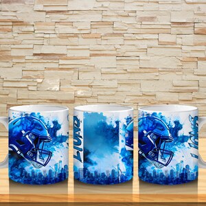 Puede incluir: Taza de cer&aacute;mica blanca con un dise&ntilde;o de casco de f&uacute;tbol americano azul y una silueta de ciudad. La taza presenta la palabra "Lions" y detalles en acuarela azul. La taza est&aacute; sobre una superficie de madera.