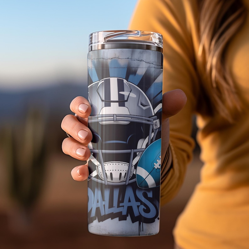 May include: A stainless steel tumbler featuring a football helmet and football graphic in shades of gray and blue. The word "DALLAS" is written in blue graffiti-style lettering. The tumbler is held by a person.
