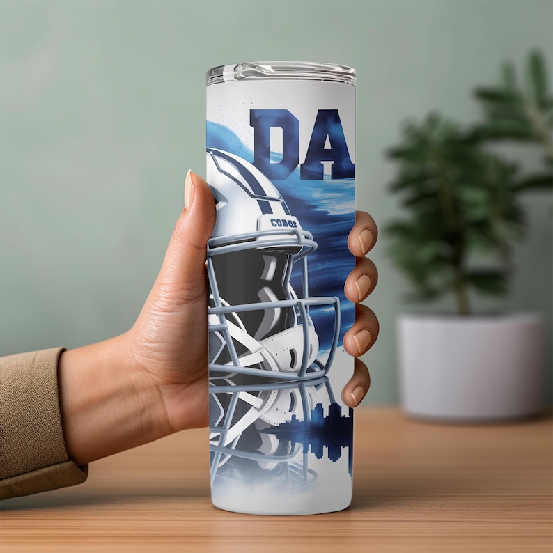 May include: A white and blue tumbler featuring a football helmet design with the letters "DA". The design includes a silhouette of a city skyline and a blue wave pattern. The tumbler is cylindrical and appears to be made of stainless steel.