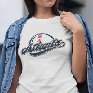 May include: White t-shirt featuring a blue and orange baseball graphic with the text "Atlanta" in a vintage style font.