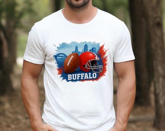May include: White t-shirt featuring a football, a red helmet, and the word "BUFFALO" in blue. The design includes a cityscape and a bridge, with red and blue paint splashes.