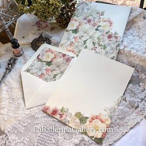 Könnte beinhalten: Briefpapier-Set mit floralen Designs. Das Set enthält ein leeres Briefpapier, einen Umschlag mit floralem Innenfutter und ein größeres Blatt mit floralem Rand. Das florale Muster enthält weiße, rosa und grüne Elemente. Die Website LaBallerinaGallery.com ist sichtbar.