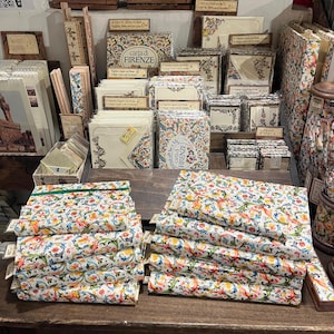 May include: A selection of stationery items with the "Carta di Firenze" design, including notebooks and notecards. The products display a vibrant floral pattern and are presented on wooden shelves. Various sizes and styles of the stationery are visible.
