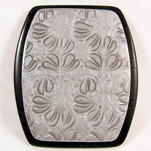 May include: A silver-colored rectangular pendant with a black border. The pendant has a raised, embossed design of a repeating floral pattern.