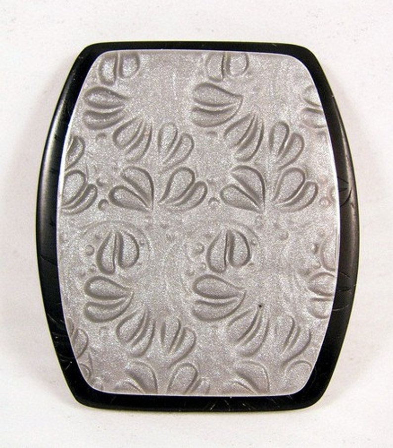 May include: A silver-colored rectangular pendant with a black border. The pendant has a raised, embossed design of a repeating floral pattern.