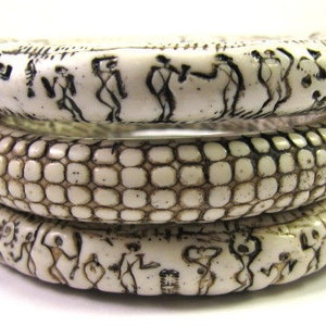 Puede incluir: Tres brazaletes de color marfil con dise&ntilde;os intrincados. El brazalete superior presenta un patr&oacute;n repetitivo de figuras, el brazalete del medio tiene un patr&oacute;n texturizado y escamoso, y el brazalete inferior presenta un patr&oacute;n repetitivo de figuras.