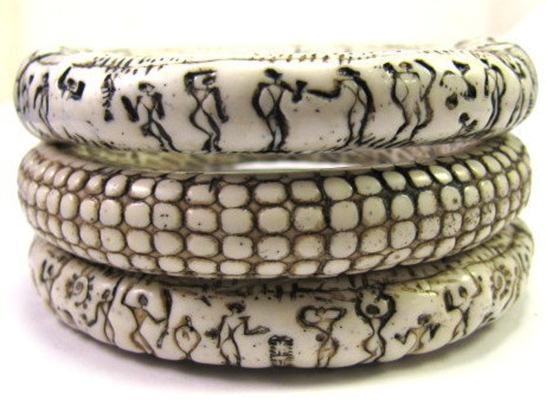 Puede incluir: Tres brazaletes de color marfil con dise&ntilde;os intrincados. El brazalete superior presenta un patr&oacute;n repetitivo de figuras, el brazalete del medio tiene un patr&oacute;n texturizado y escamoso, y el brazalete inferior presenta un patr&oacute;n repetitivo de figuras.
