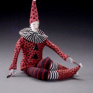 May include: A red and black polka dot clown doll with a white collar and a black and white striped hat. The doll is sitting with its legs crossed and is holding a small stick in its hand.