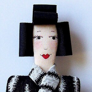 May include: A handmade doll with black hair, a black hat, and a white and black patterned dress. The doll has a painted face with red lips.