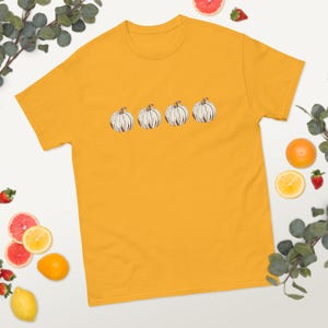 May include: Mustard yellow t-shirt featuring a row of four white pumpkins with black outlines. The pumpkins have green stems. The shirt is made of a soft material. The shirt is a casual wear item.