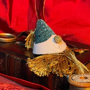May include: A vintage book with a green and white hat with a gold tassel on top. The book is on a red fabric surface.