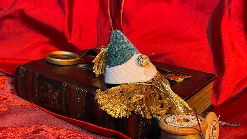 May include: A vintage book with a green and white hat with a gold tassel on top. The book is on a red fabric surface.