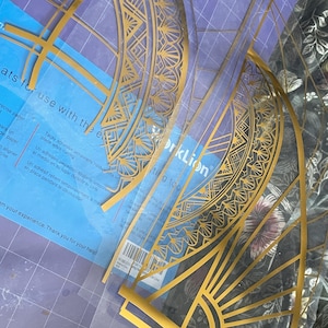 Puede incluir: Plantillas doradas de estilo Art D&eacute;co sobre una esterilla de corte azul. Las plantillas est&aacute;n hechas de un material fino y transparente y presentan intrincados dise&ntilde;os geom&eacute;tricos. El embalaje incluye texto sobre adhesivo temporal y herramientas de corte.