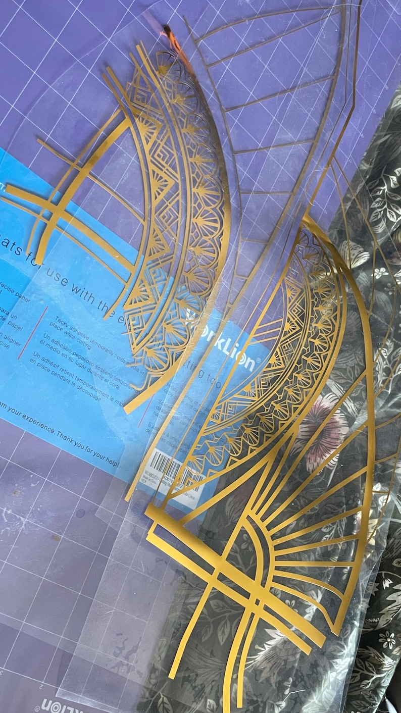 Puede incluir: Plantillas doradas de estilo Art D&eacute;co sobre una esterilla de corte azul. Las plantillas est&aacute;n hechas de un material fino y transparente y presentan intrincados dise&ntilde;os geom&eacute;tricos. El embalaje incluye texto sobre adhesivo temporal y herramientas de corte.